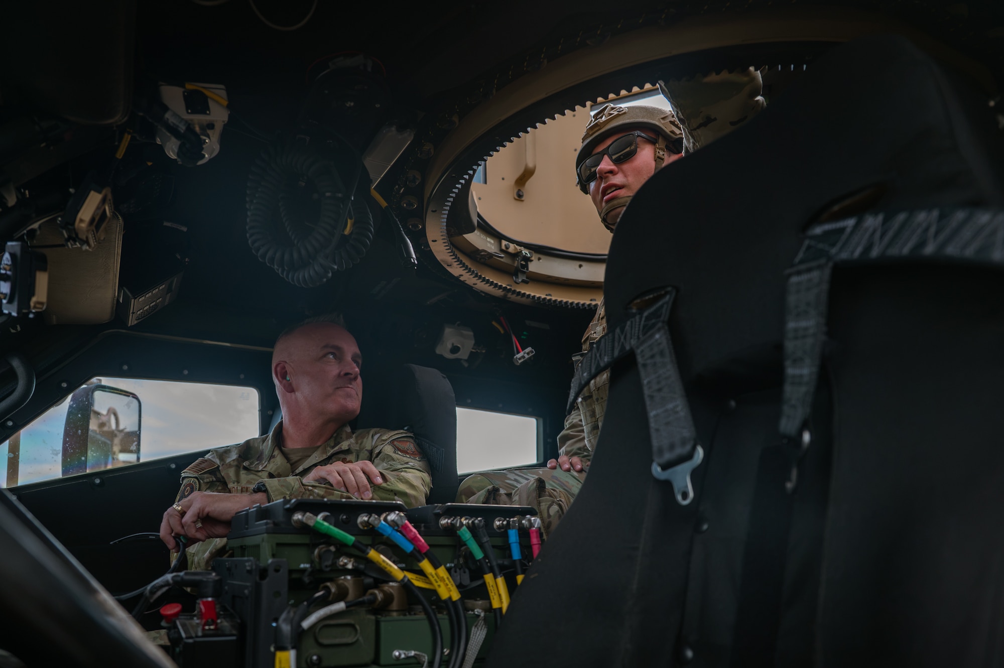 A man in military uniform sits in the passenger seat of a convoy vehicle as another man in military uniform and helmet stands in the hatch area looking toward the sky.