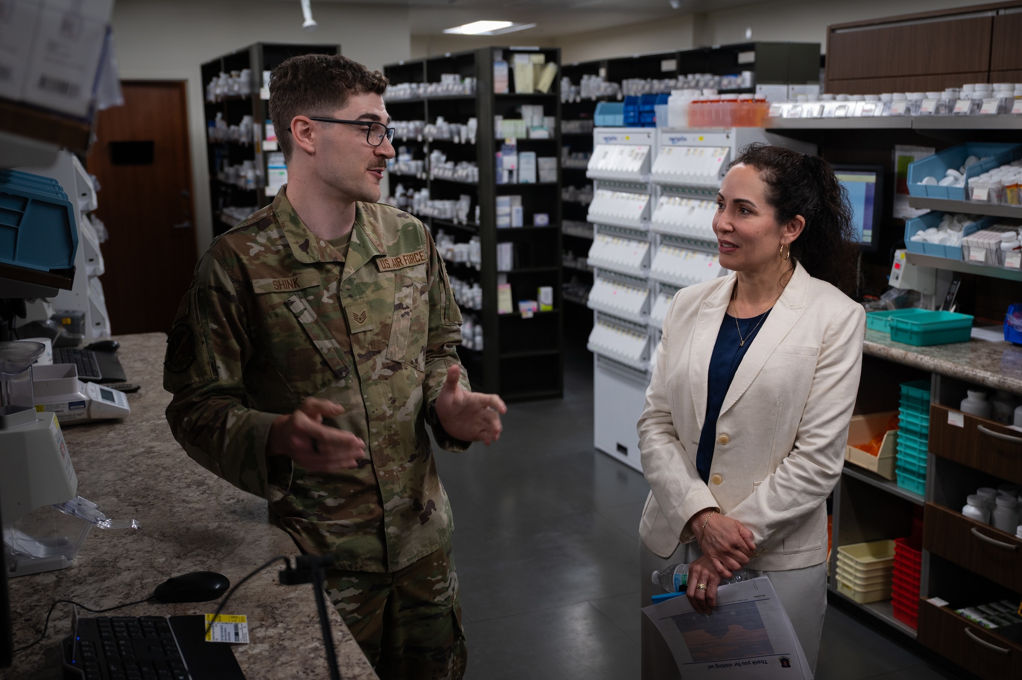 A man in camouflage military uniform stands in front of a woman wearing a white blazer as they talk.