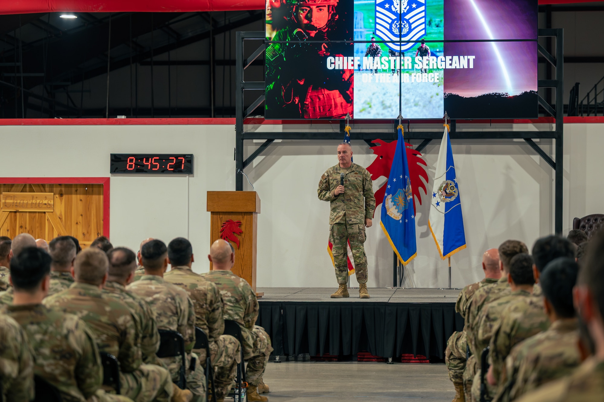 A man in military camouflage uniform stands on stage in front of a crowd of other uniformed military members.