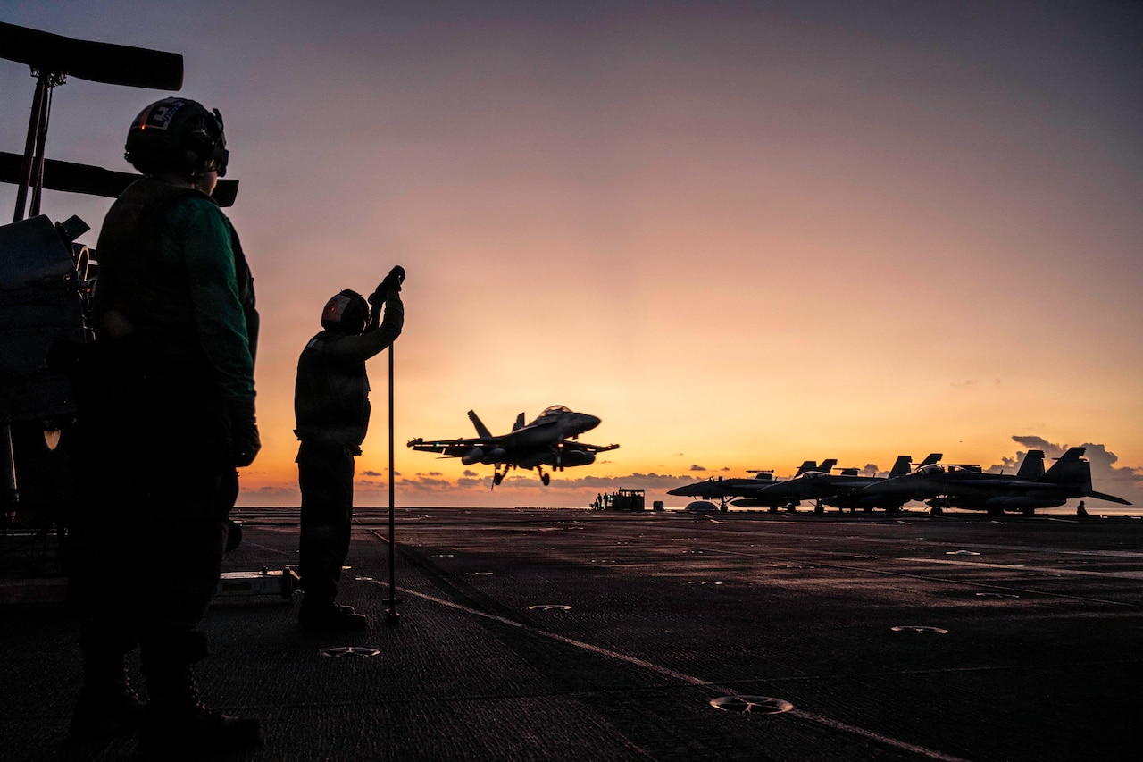 A military jet lands on the deck of an aircraft carrier at sunset; there are silhouettes of two people in military uniforms standing on the ship observing in the foreground.