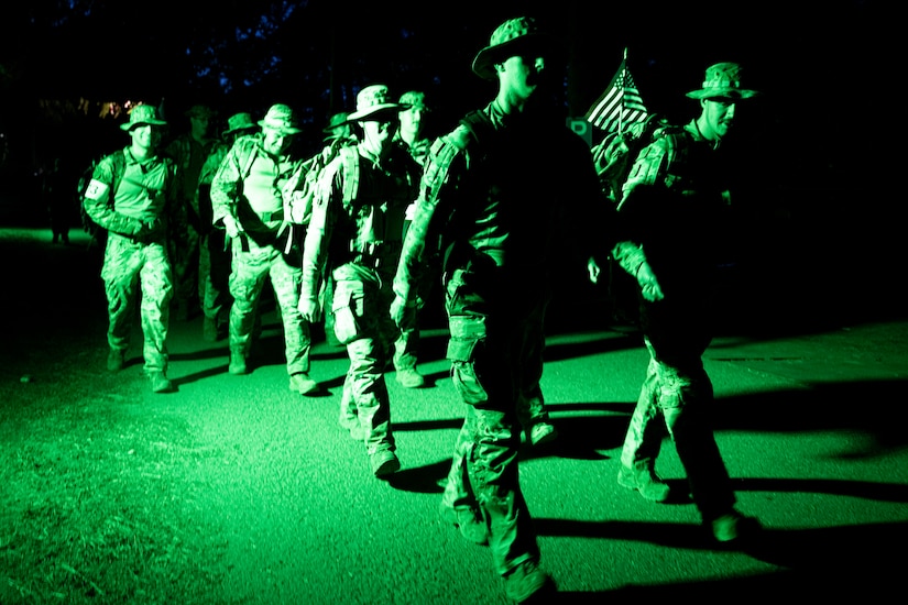 A group of people wearing camouflage military uniforms march in the dark, illuminated by green light.