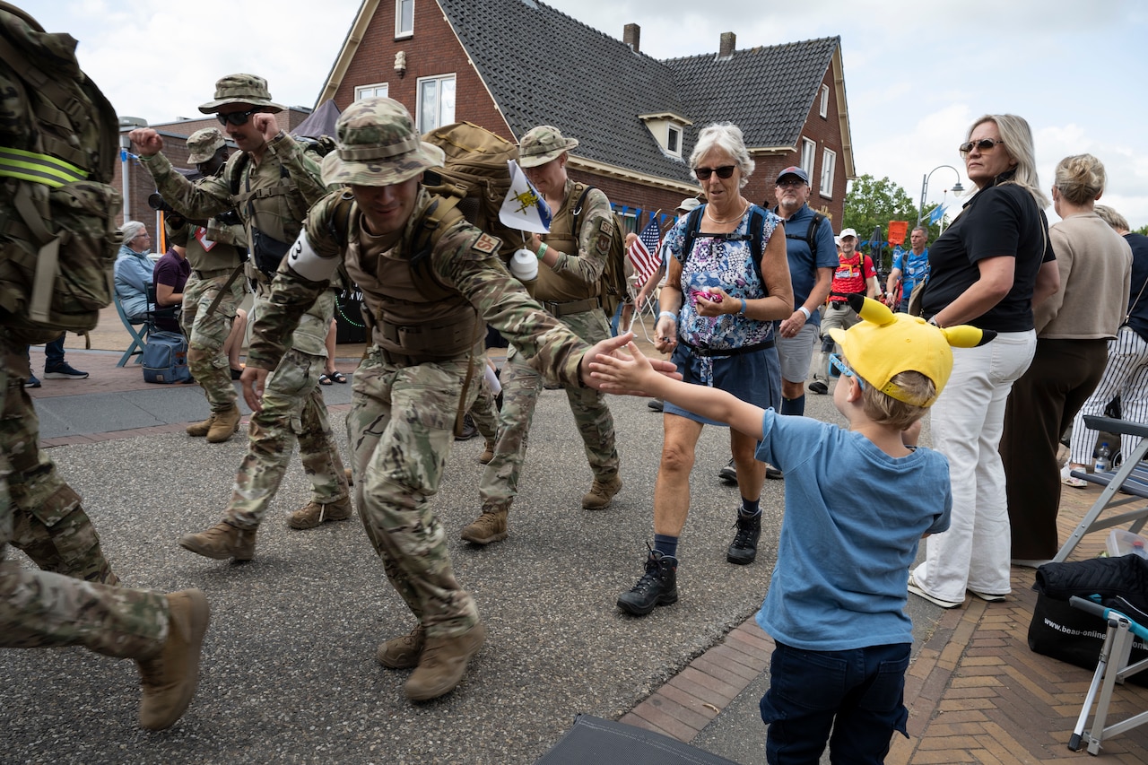 A man wearing a camouflage military uniform high-fives a boy as he marches along a city route with other people in similar attire.