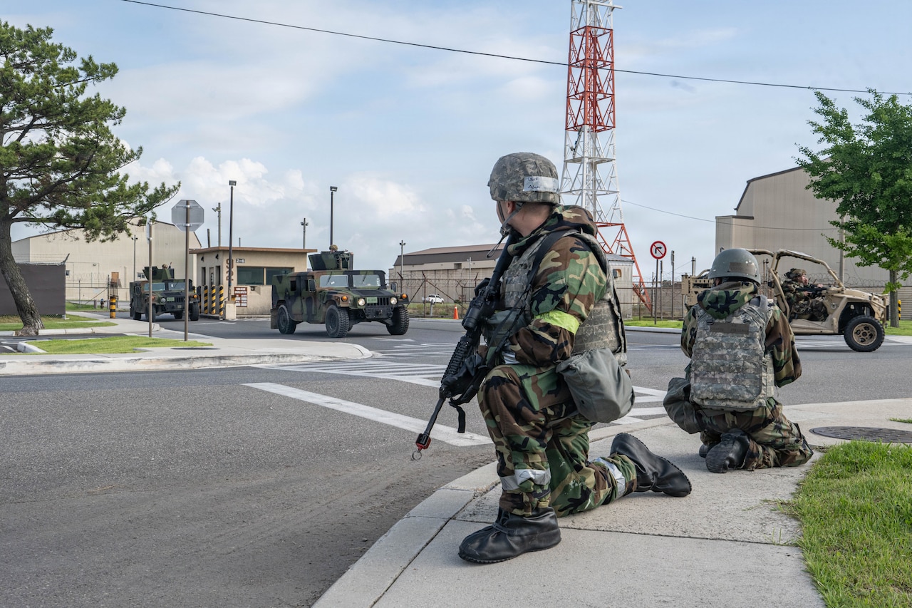 Two people in camouflage military uniforms and tactical gear kneel on a sidewalk while providing security for other military personnel coming onto the base.