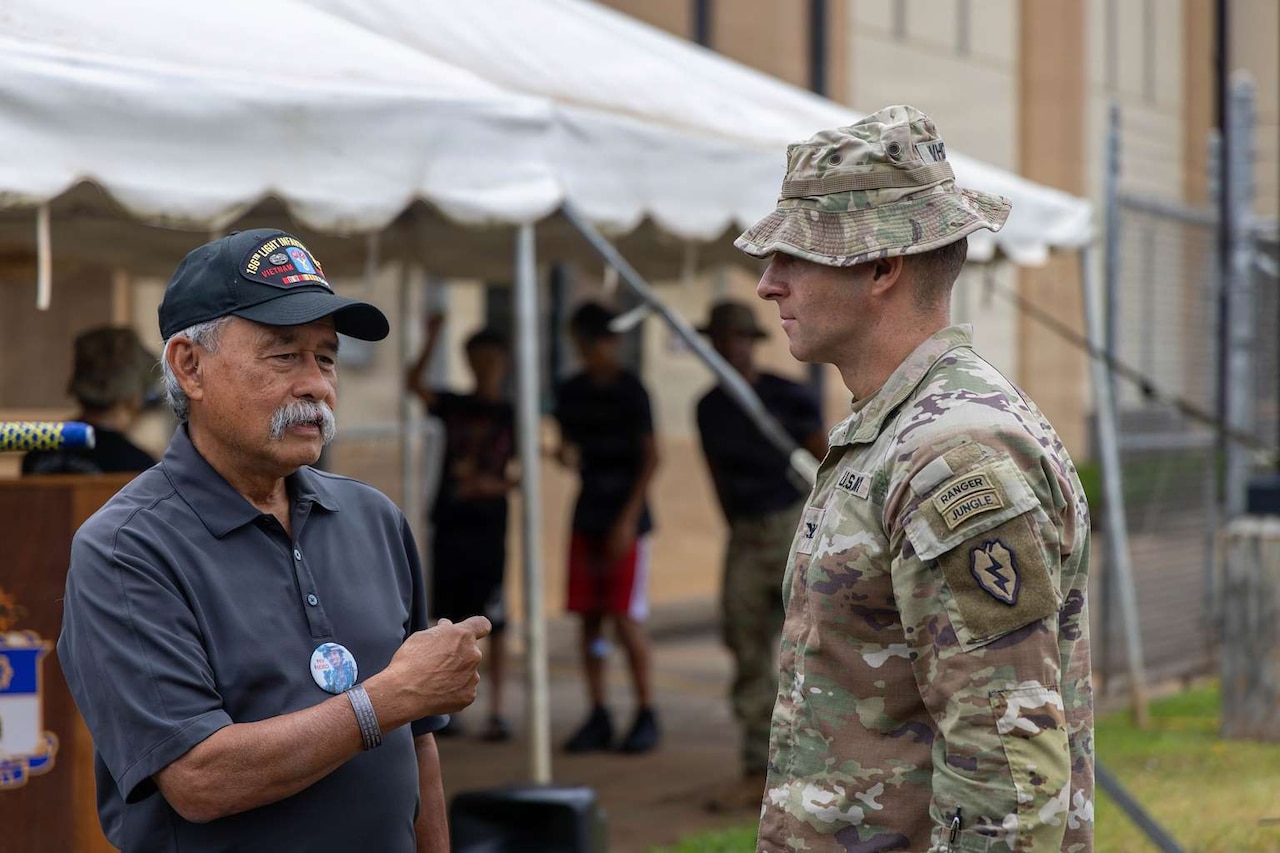 A man in casual attire speaks to a man in a military camouflage uniform outside.