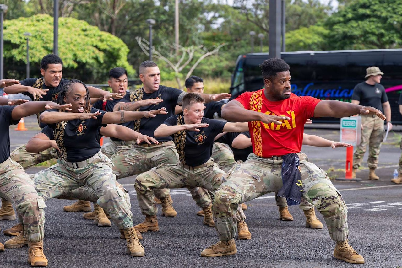 Nearly a dozen people in T-shirts and camouflage military pants crouch in a fighting stance outside with trees in the background.
