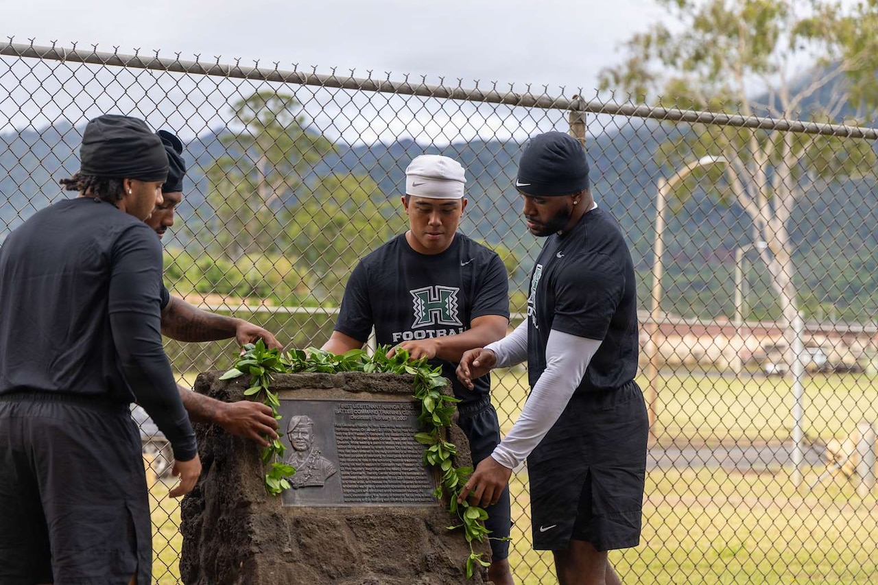 Four men in athletic attire place a Hawaiian lei at a memorial outside.