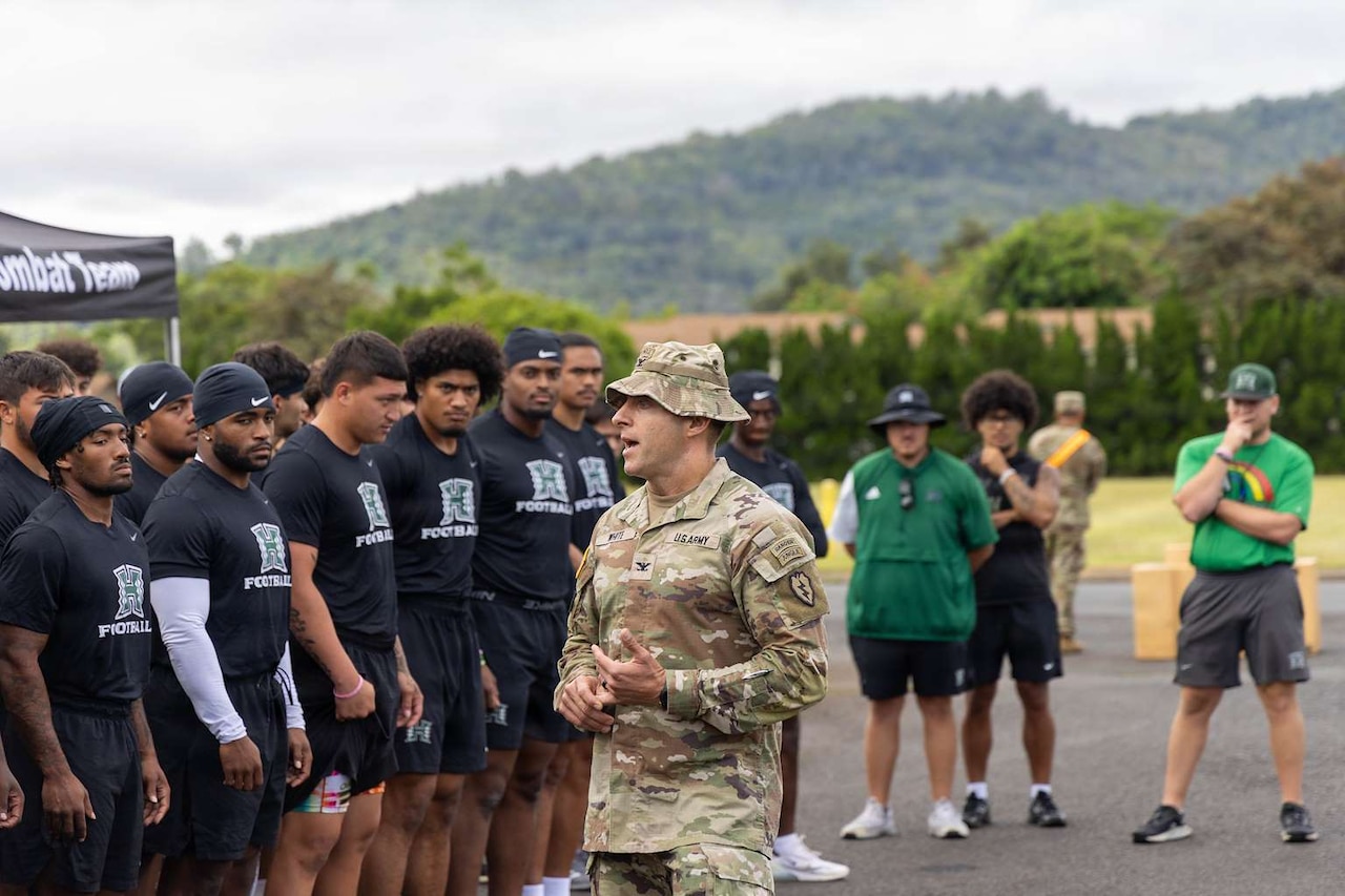 A man in a camouflage military uniform speaks to more than a dozen people in athletic attire outside with mountains and trees in the background.