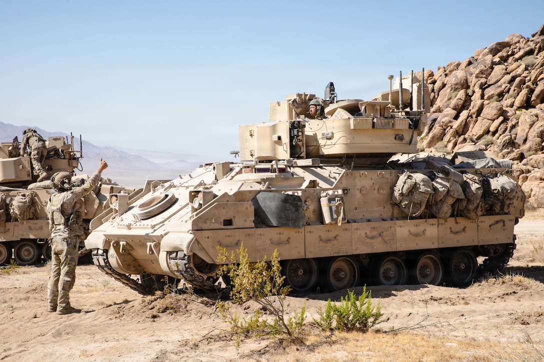 A soldier gives a thumbs-up while standing in front of a soldier operating a tank on a dirt road in front of a rocky cliff.