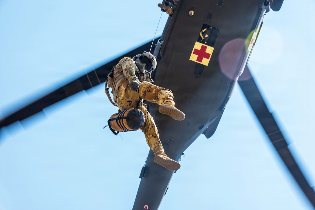 A soldier wearing protective face and head gear is hoisted to an airborne helicopter as seen from below against a blue sky.