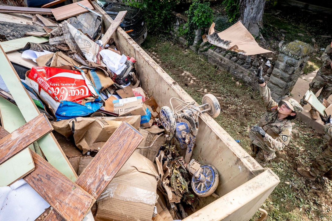 A National Guardsman throws debris into a large container of trash near a wooded area.