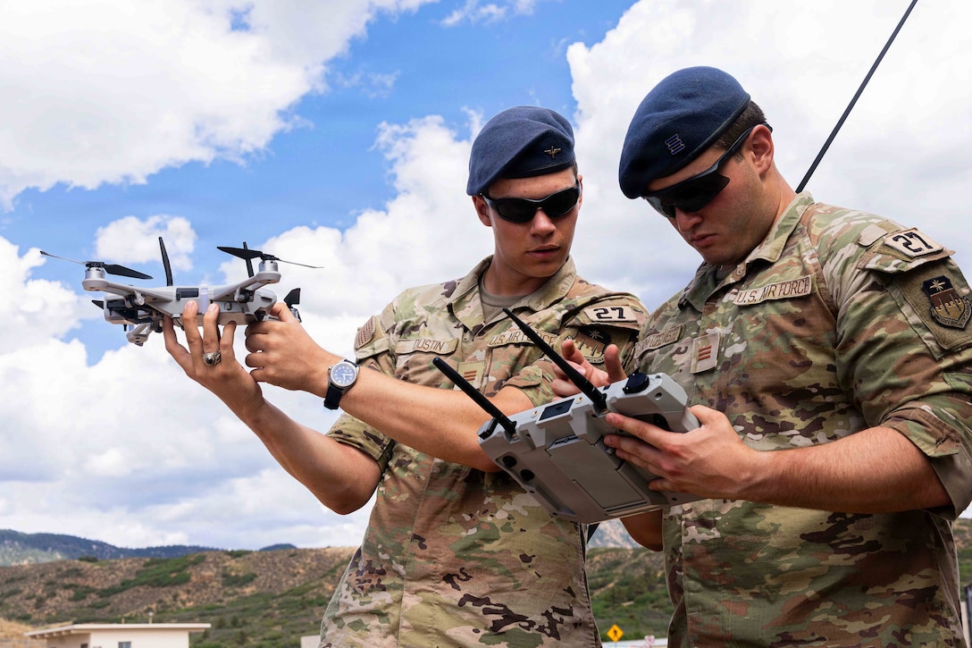 One Air Force cadet holds a remote as the other holds a drone toward a blue sky with mountains in the background.