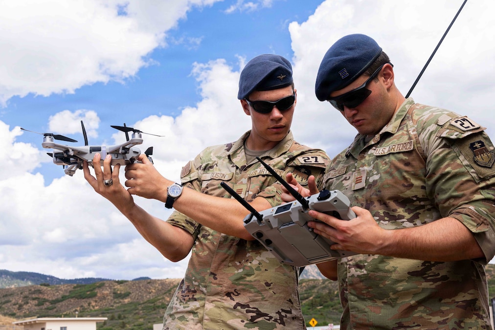 One Air Force cadet holds a remote as the other holds a drone toward a blue sky with mountains in the background.