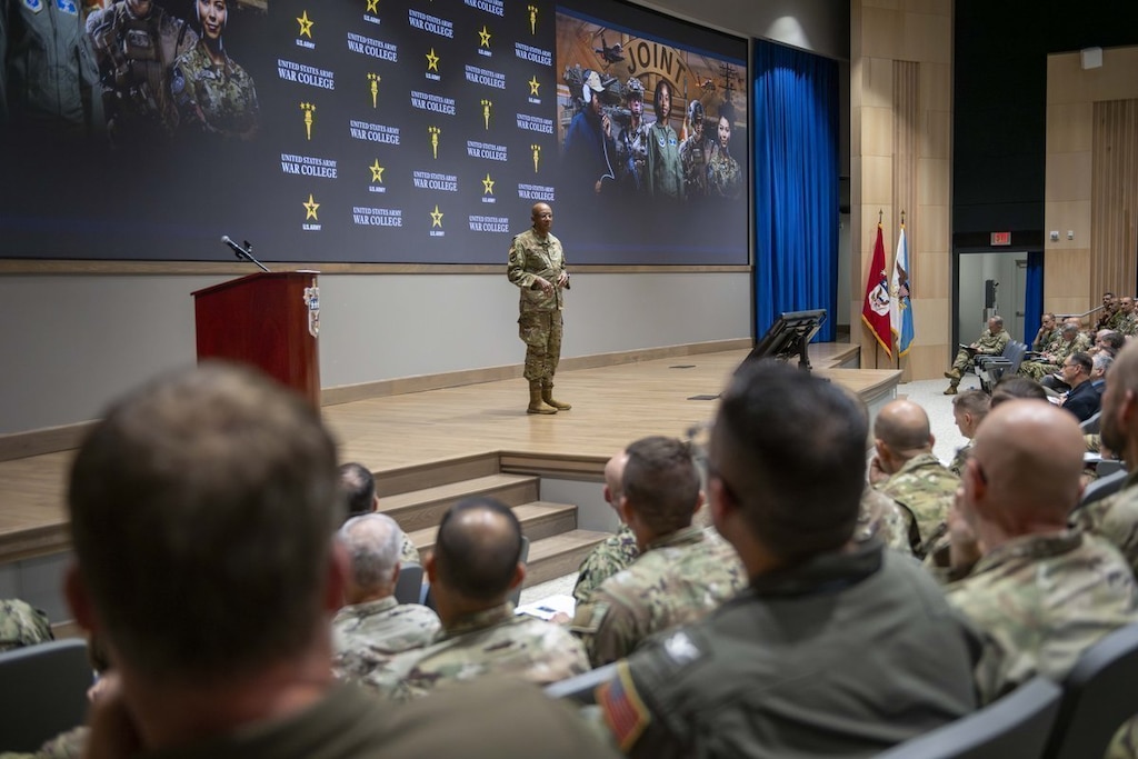 General Charles Q. Brown, Jr., Chairman of the Joint Chiefs of Staff, stands on stage addressing a large hall of senior military students at the U.S. Army War College.