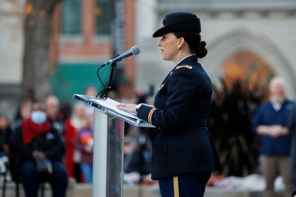 An officer in Class A dress uniform speaks from a podium to a large outdoor assembly during a military ceremony.