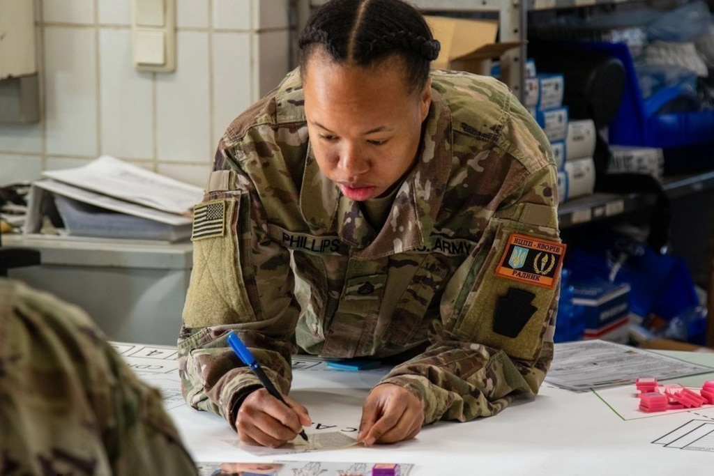 A female service member in utility uniform uses a marker to plot coordinates on a large tabletop wargame map.