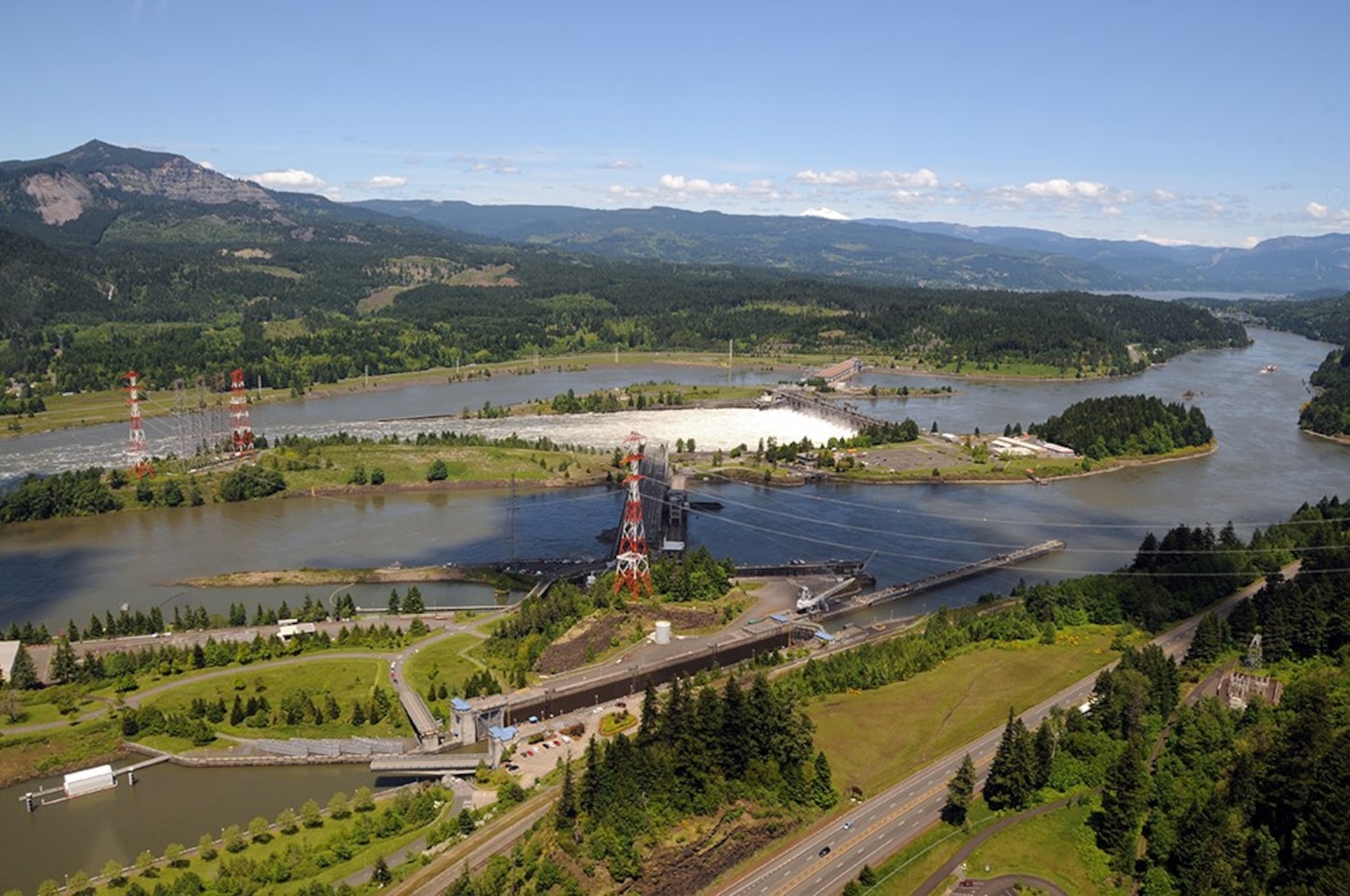 An aerial, wide-angle photograph of the Bonneville Lock and Dam on the Columbia River on a sunny day. The image captures the powerhouse and the spillway, which is actively releasing white, turbulent water. In the foreground, navigation locks and a highway are visible, set against a landscape of green, forested hills that stretch into the mountainous background of the Columbia River Gorge under a clear blue sky.