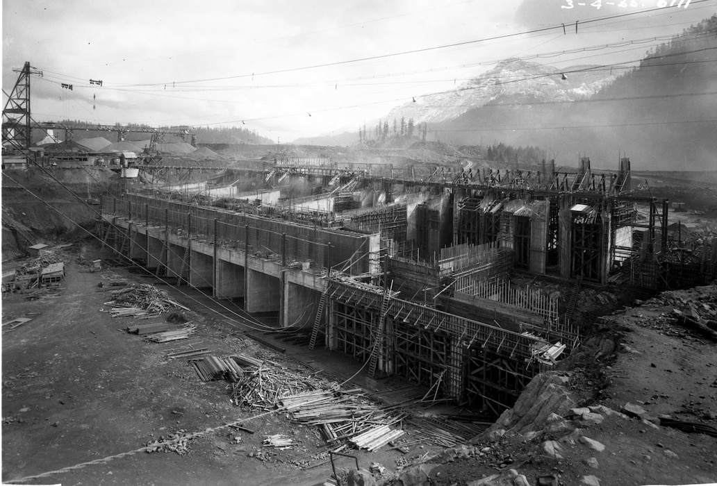 A wide-angle, black-and-white historical photograph from 1935 showing the Bonneville Dam during its construction. The vast concrete structure is covered in extensive wooden scaffolding and formwork, with exposed rebar visible throughout. The foreground is a dirt construction site with piles of lumber and materials, while the steep, forested mountains of the Columbia River Gorge rise in the background.