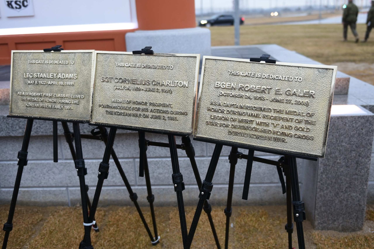 Three plaques, each dedicated to service members, are displayed on easels outside during a ceremony.