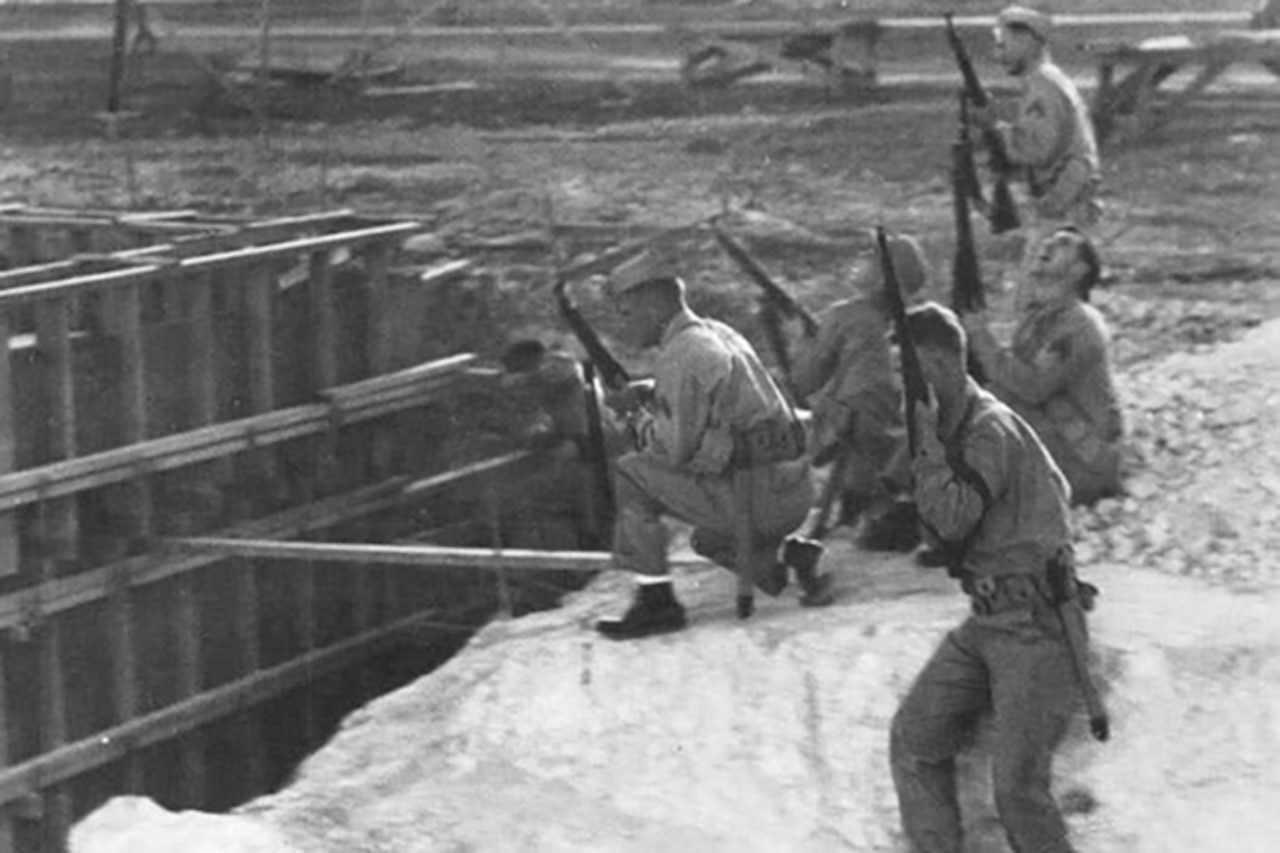 Five men in World War II-era military uniforms crouch and stand outside as they point their rifles toward the sky.