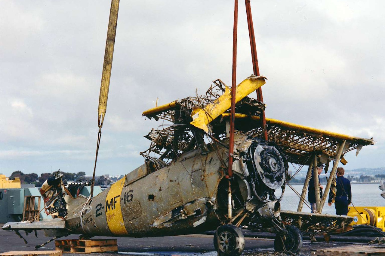 A wrecked World War II-era plane is lowered to the ground near an ocean.