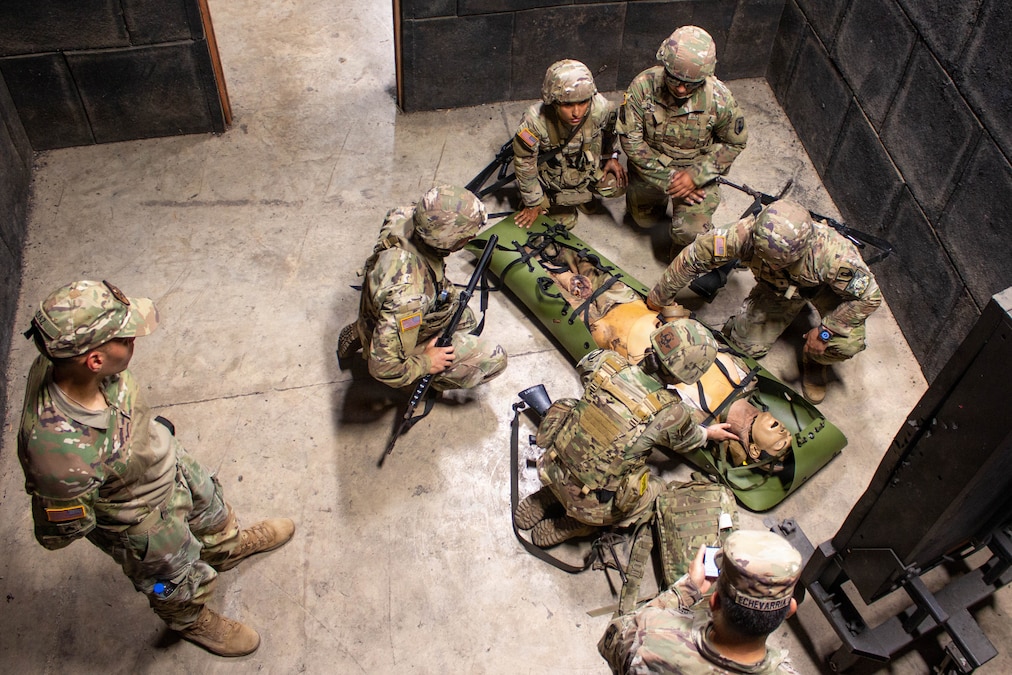 Five soldiers wearing camouflage uniforms, helmets and carrying rifles kneel around a simulated casualty mannequin strapped into a medical evacuation litter while two other soldiers, also wearing camouflage uniforms and hats, stand and observe.