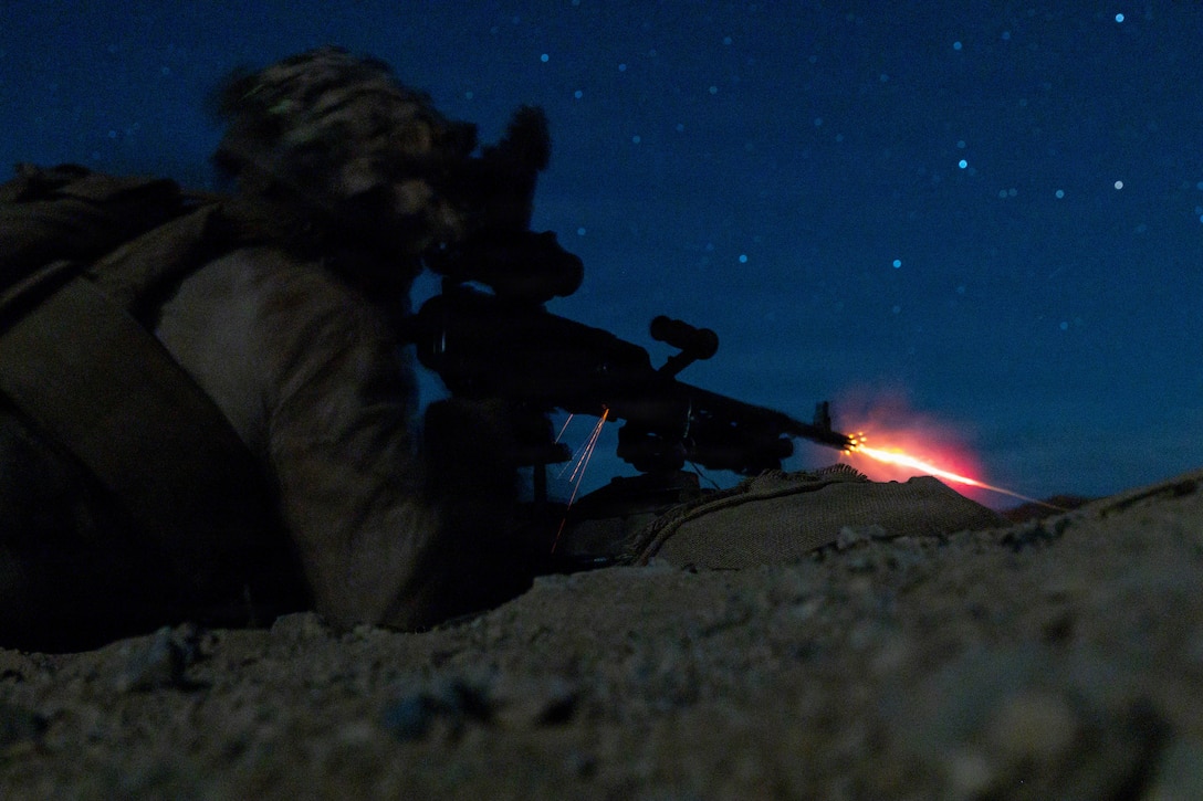 Flames burst from the muzzle of a machine gun fired by a Marine lying in the prone position, looking through a night vision device at night, with stars visible in the sky.