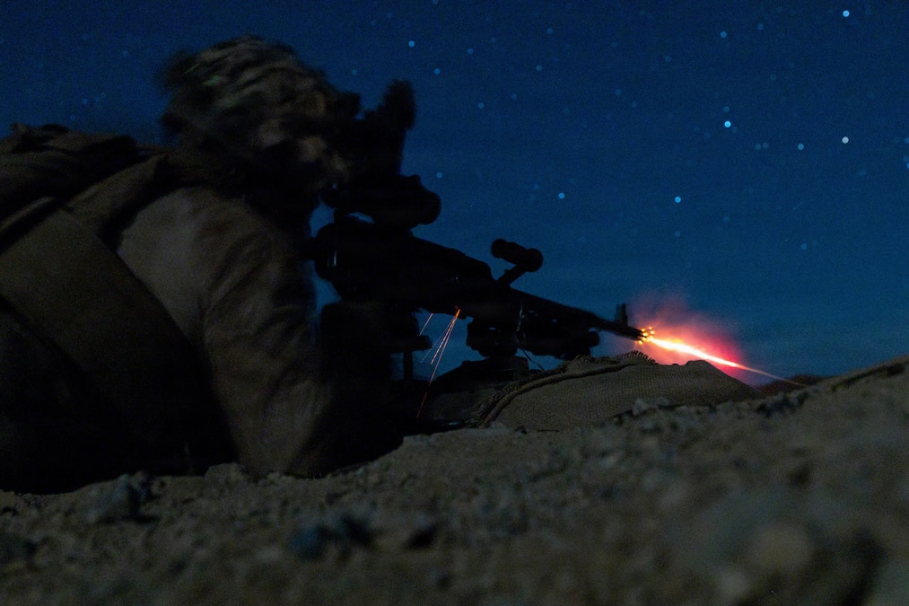 Flames burst from the muzzle of a machine gun fired by a Marine lying in the prone position, looking through a night vision device at night, with stars visible in the sky.