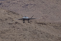 A U.S. Air Force YFQ-42A Dark Merlin aircraft is descending from the sky with mountain terrain in the background.