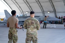 Two U.S. Airmen stand with their backs turned to the camera observing the  YFQ-44A Fury aircraft inside an open hangar.