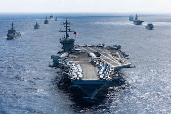 An aircraft carrier sails in formation with eight other military ships of various types and sizes sailing behind it.