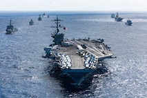 An aircraft carrier sails in formation with eight other military ships of various types and sizes sailing behind it.