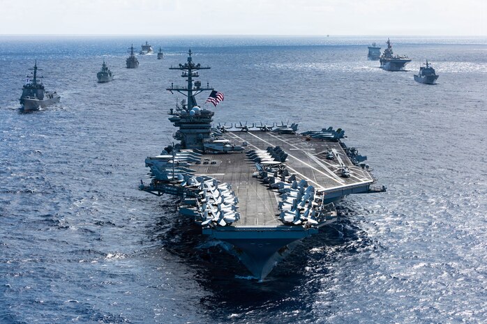 An aircraft carrier sails in formation with eight other military ships of various types and sizes sailing behind it.