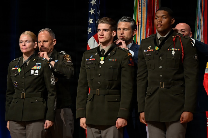 A woman and two men wearing military dress uniforms stand facing forward in a line while three men behind them place medals around their necks.