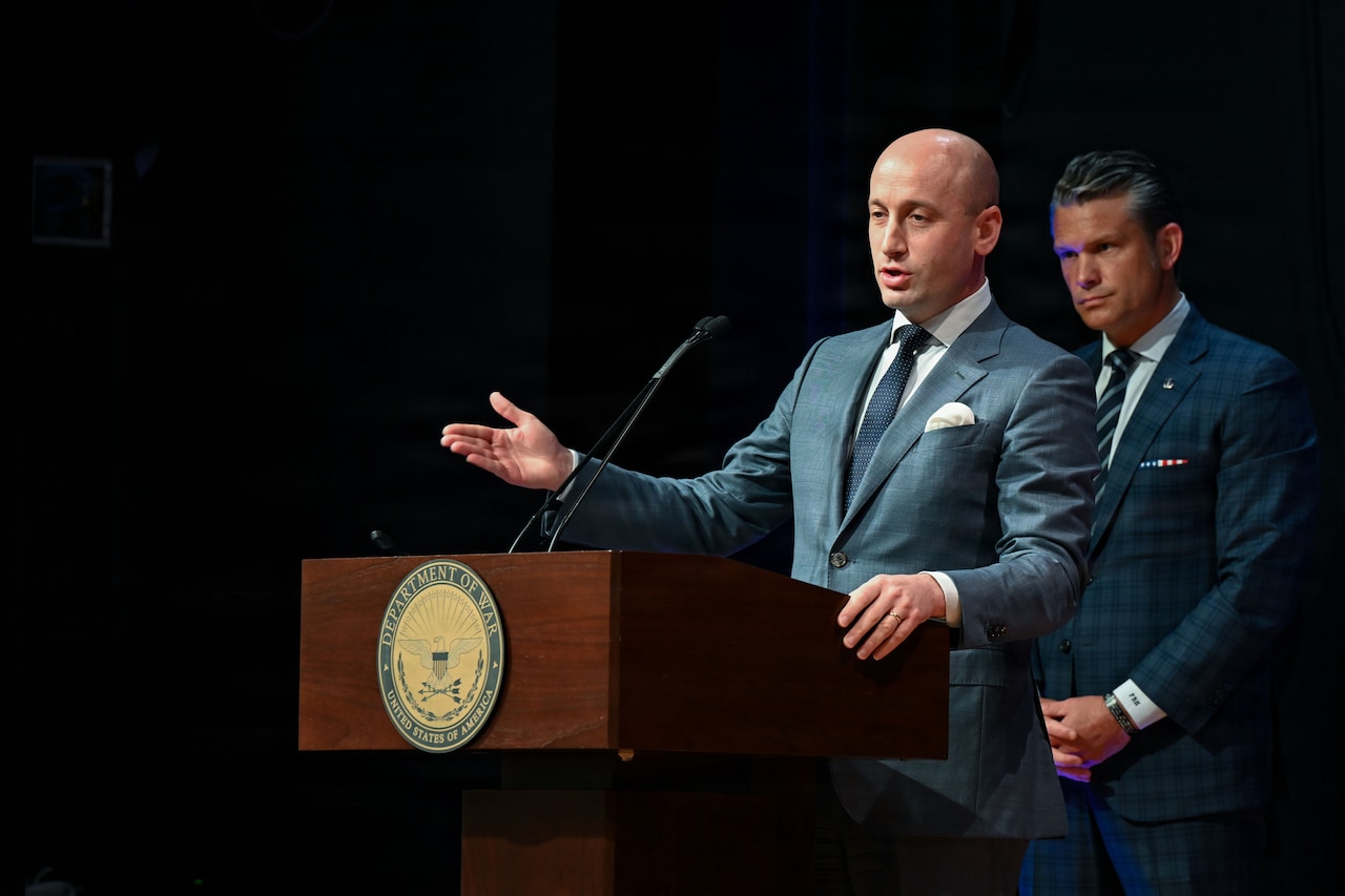 A man stands behind a lectern. Another man stands behind him.