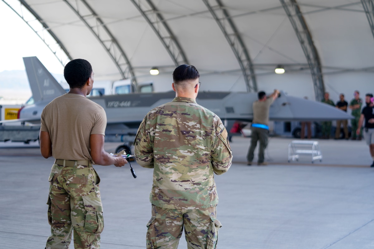 Two U.S. Airmen stand with their backs turned to the camera observing the  YFQ-44A Fury aircraft inside an open hangar.