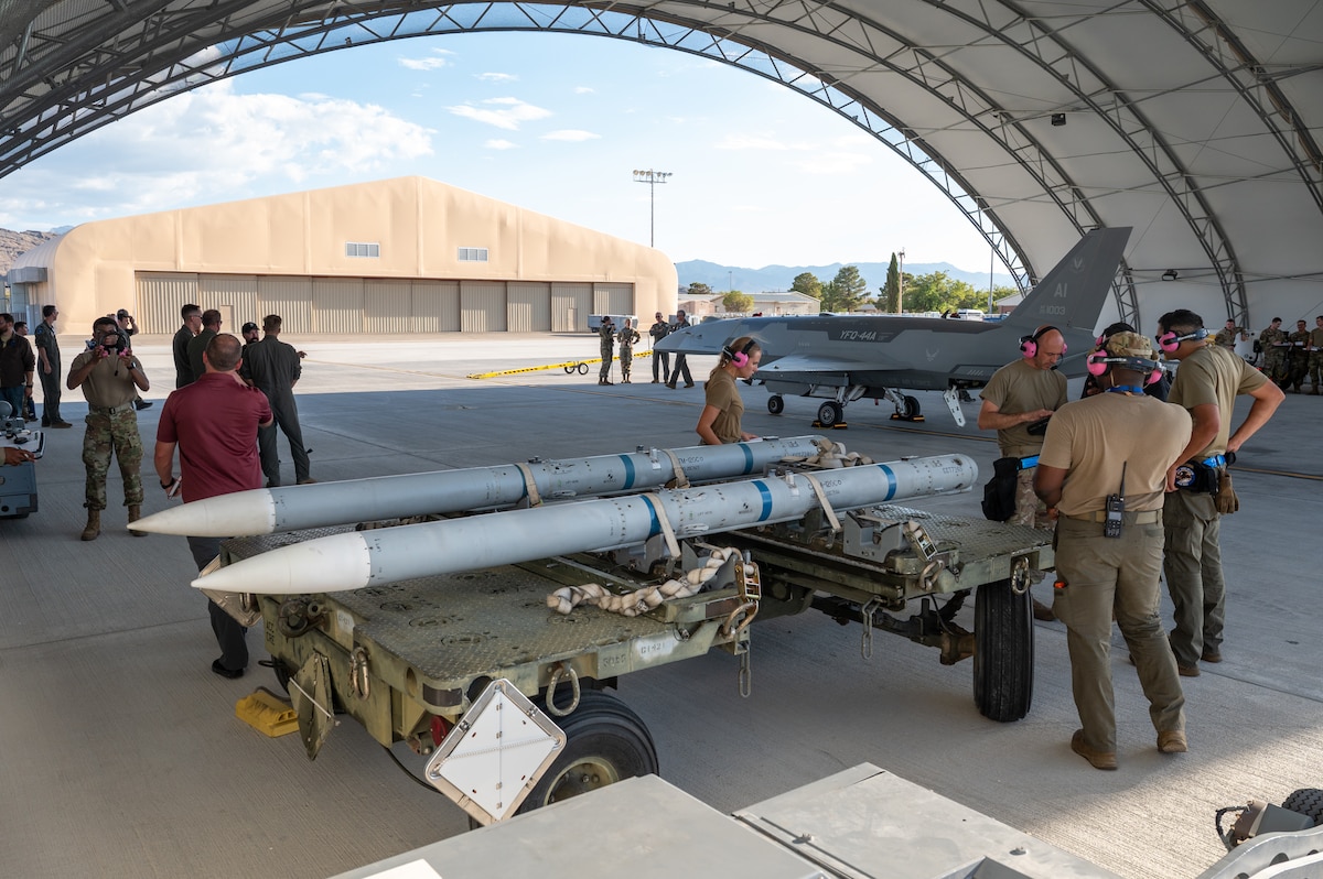 Airmen gather below a hangar with a YFQ-44A Fury aircraft and missiles in the vicinity.