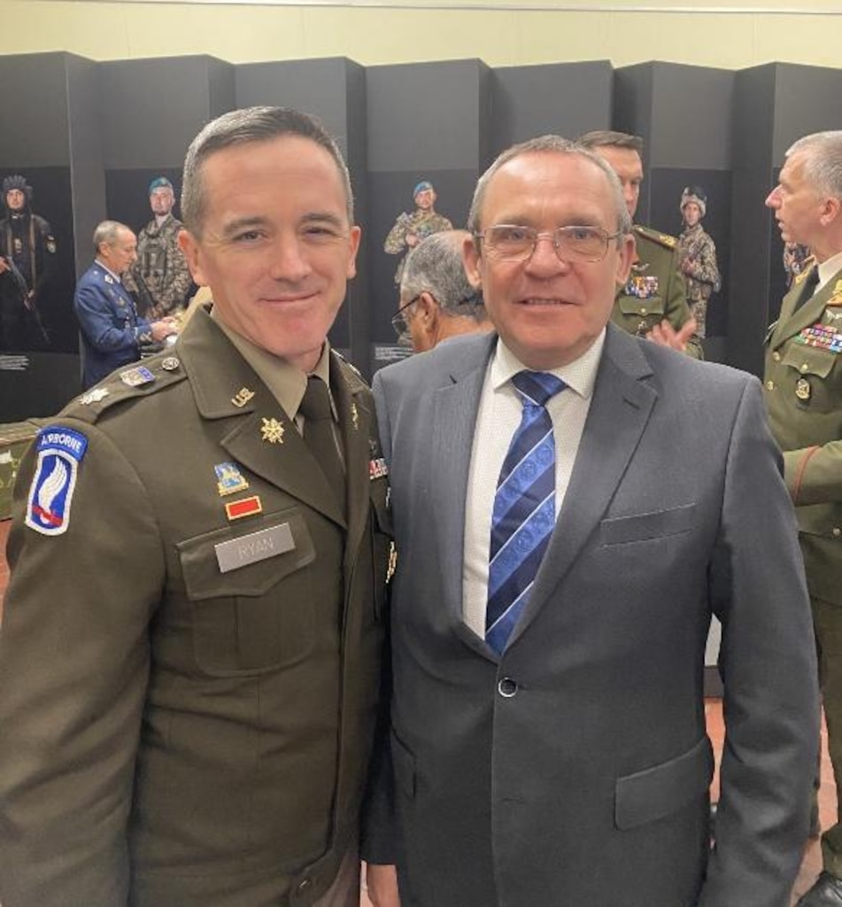 A military officer in Class A uniform and a civilian in a suit and tie standing together.