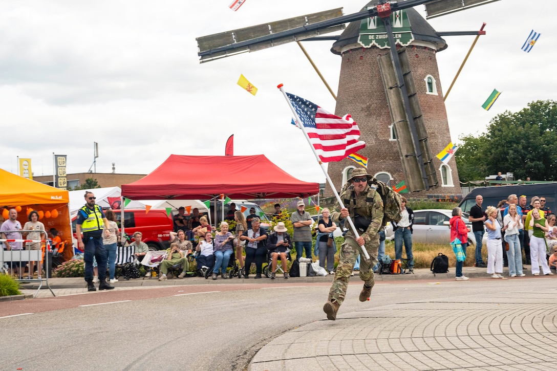 An airman in a camouflage uniform runs with an American flag in front of a tower as seated and standing spectators cheer from the sidelines.