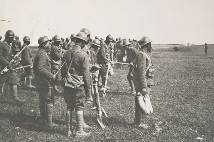 More than a dozen Black men in World War I-era uniforms and helmets hold weapons outside in a grassy field.