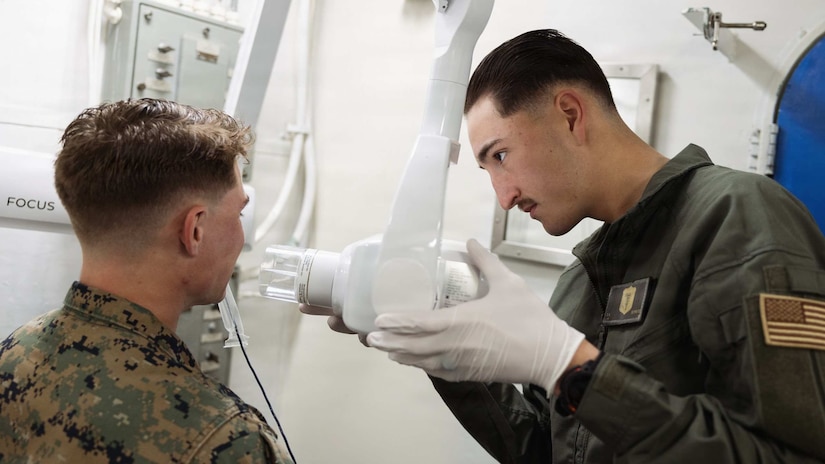 A man wearing casual attire adjusts a medical device on the face of a seated man wearing a camouflage military uniform.