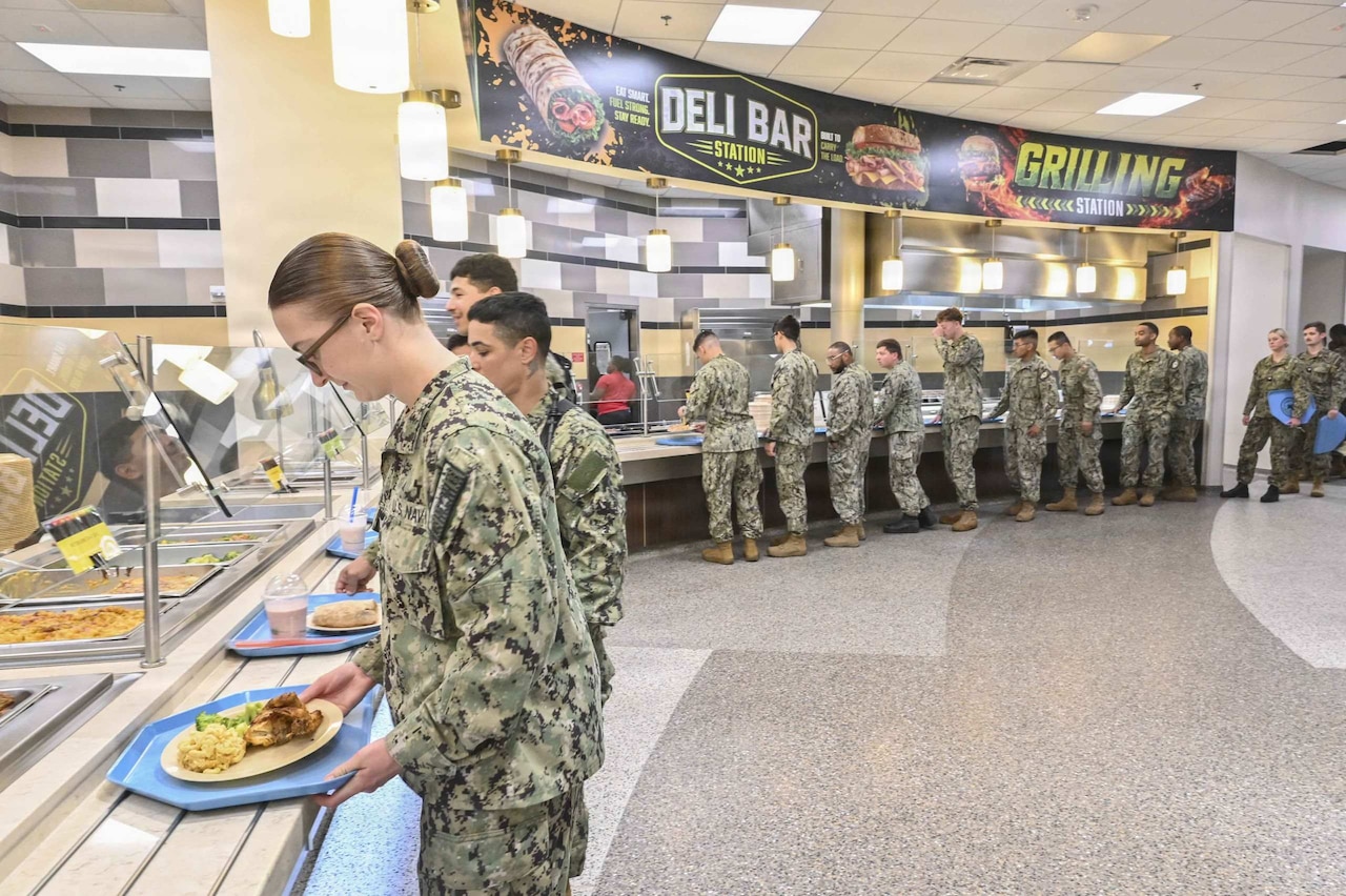 More than a dozen people wearing camouflage military uniforms stand in line at a dining facility with trays of food.