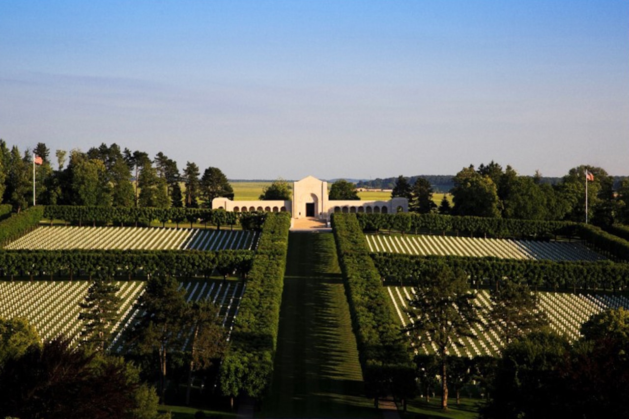 An aerial view of a cemetery with thousands of tombstones, trees and greenery under a blue sky; American flags wave from poles in the distance.