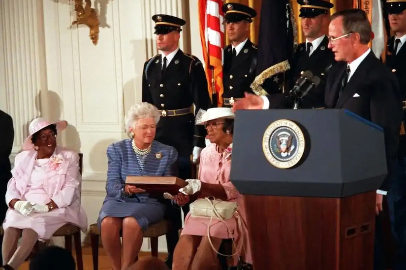 A man in business attire stands at a lectern and speaks into a microphone as three women sit in chairs. The middle woman hands a wooden award case to the woman on her left; four men in formal military uniforms stand at attention in the background.