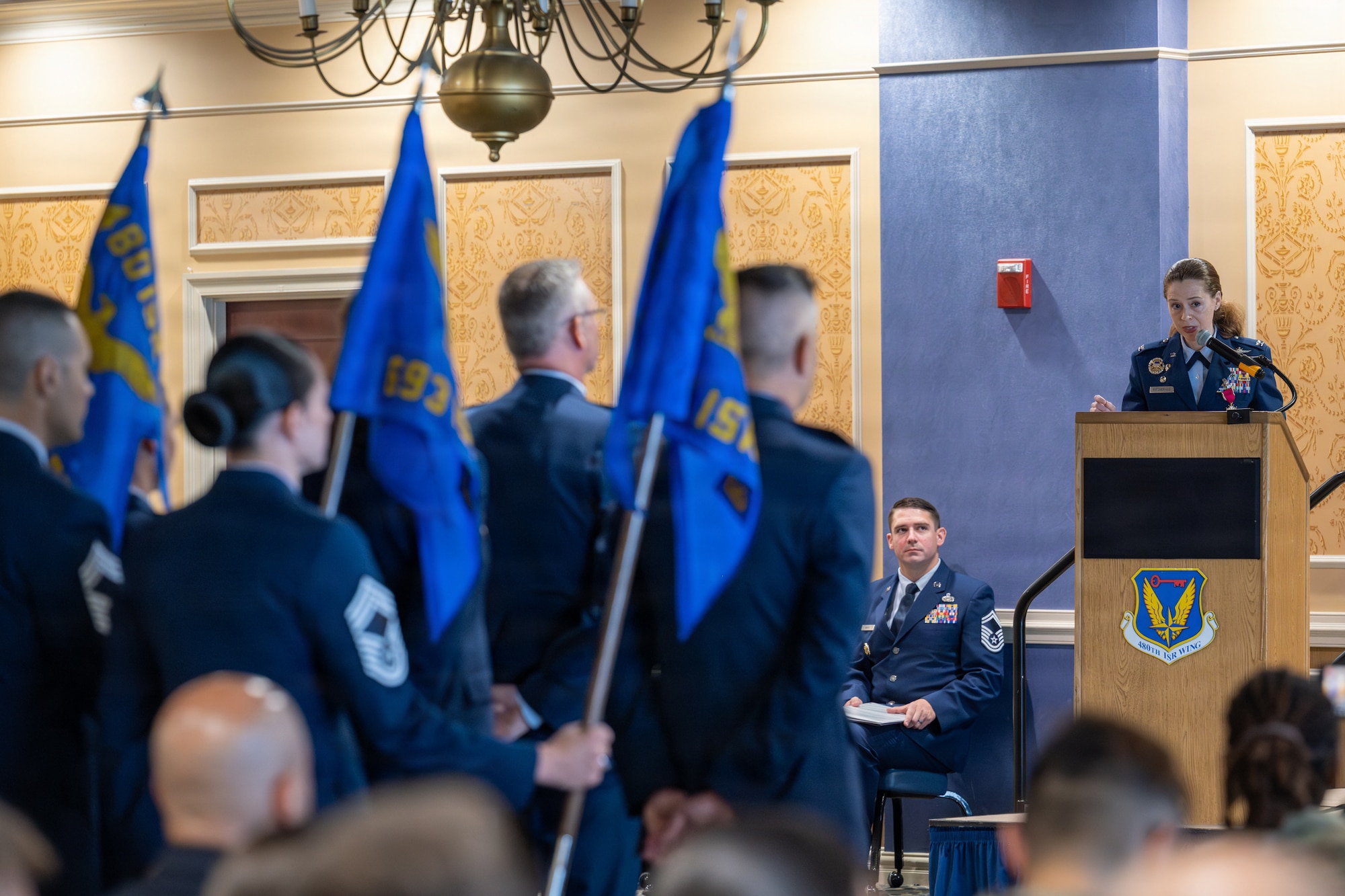 A group of Airmen stand in front of an Airman at a podium.
