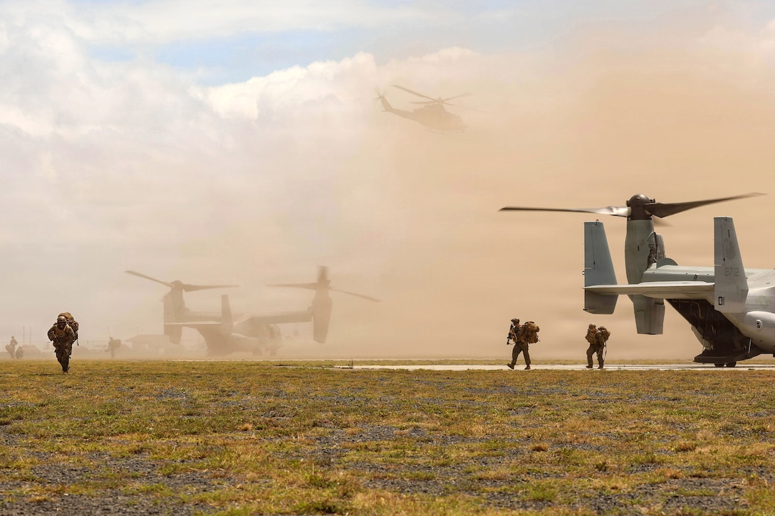 Marines walk off a runway where two Osprey aircraft are sitting and onto grass, as dust hangs in the air. A helicopter flies above clouds in the background.