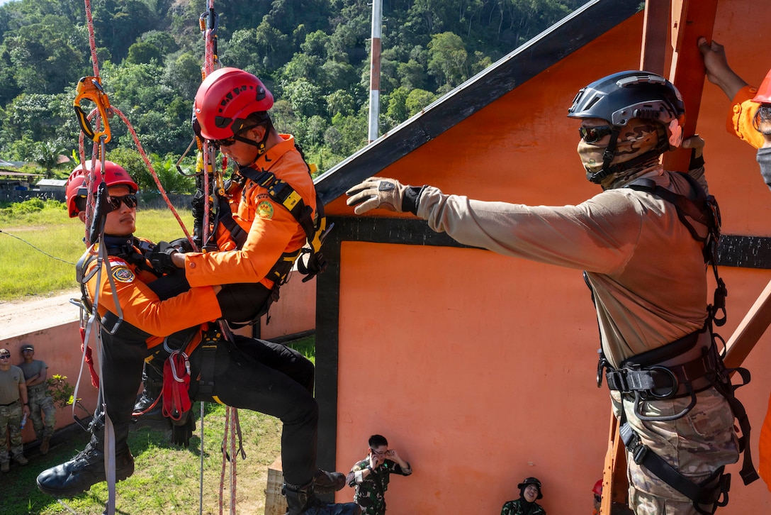 A soldier in a camouflage uniform and mask instructs two Indonesian service members. One of the service members is sitting in a sling in the air while the other is sitting in their lap.
