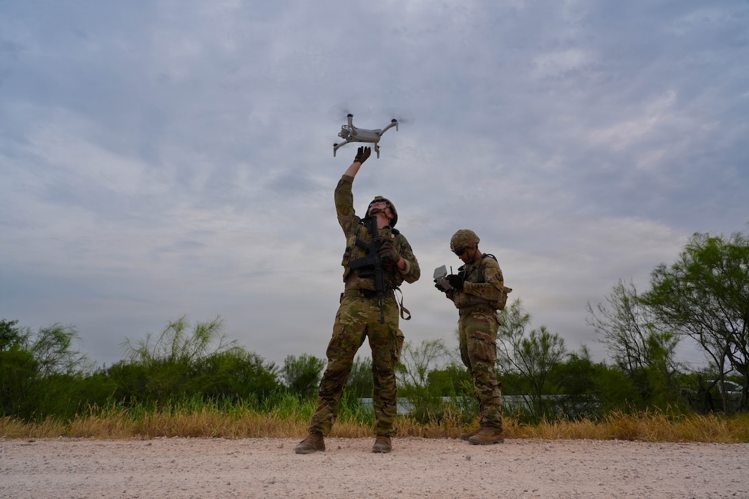 A soldier reaches up to retrieve a small drone while another soldier looks at a small device in their hands. They stand on a rocky road, with weeds and a cloudy sky in the background.