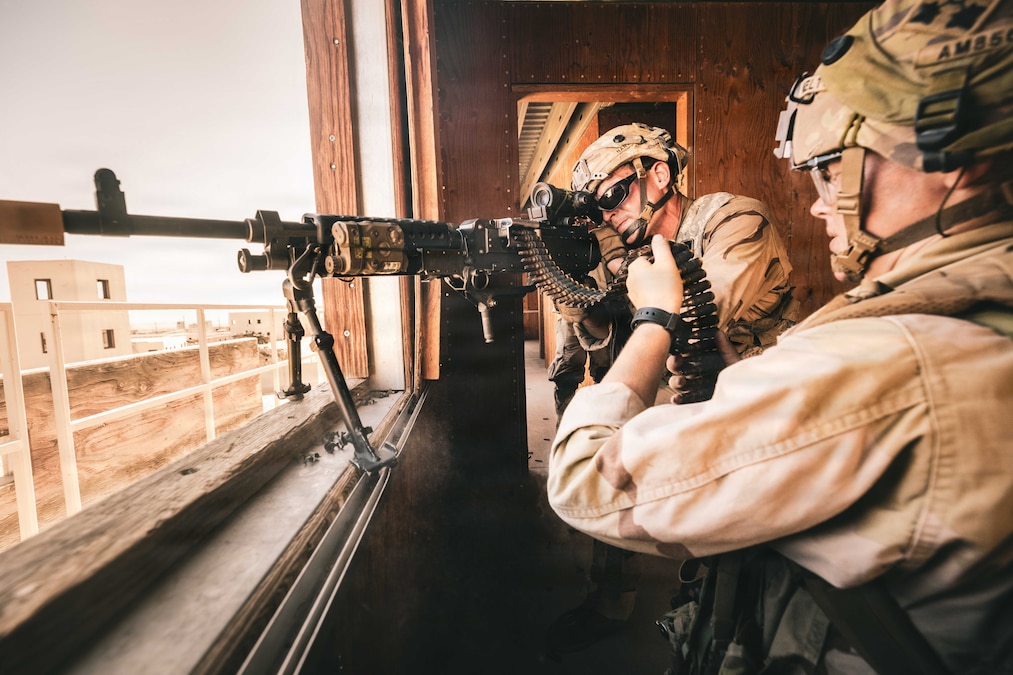 A soldier aims down the sights of a rifle pointed out a window, while another soldier holds ammunition.