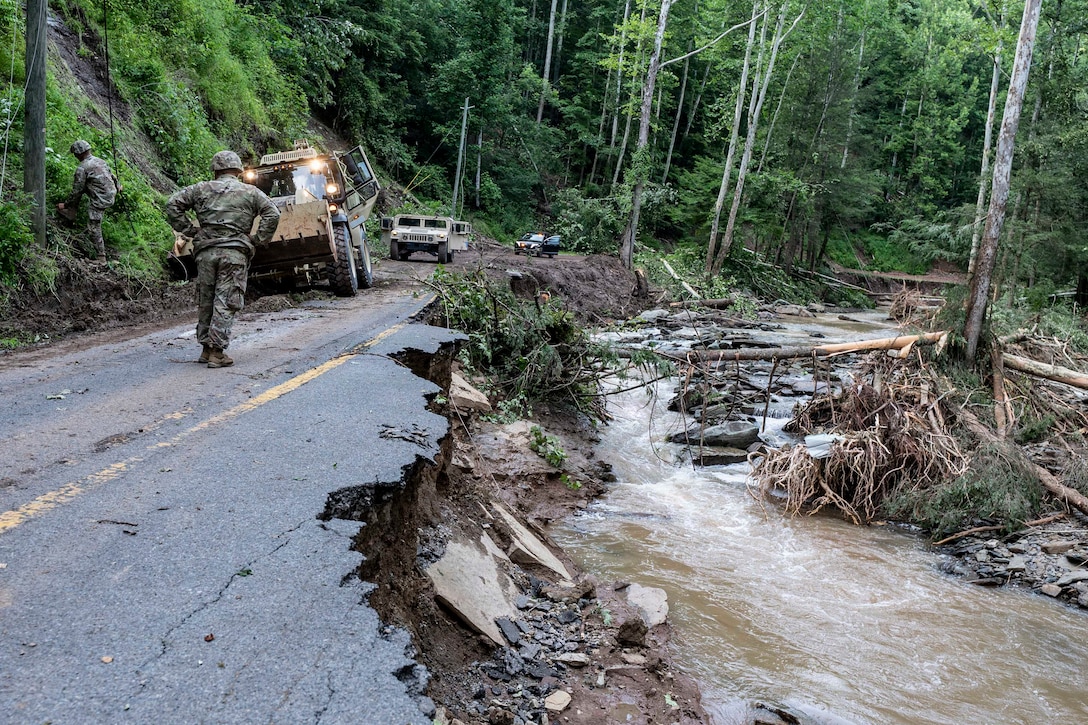 A person in a camouflage military uniform stands in front of a parked bulldozer on a partially destroyed road as another person in similar attire stands on a nearby hill; trees, water and mud surround the road.