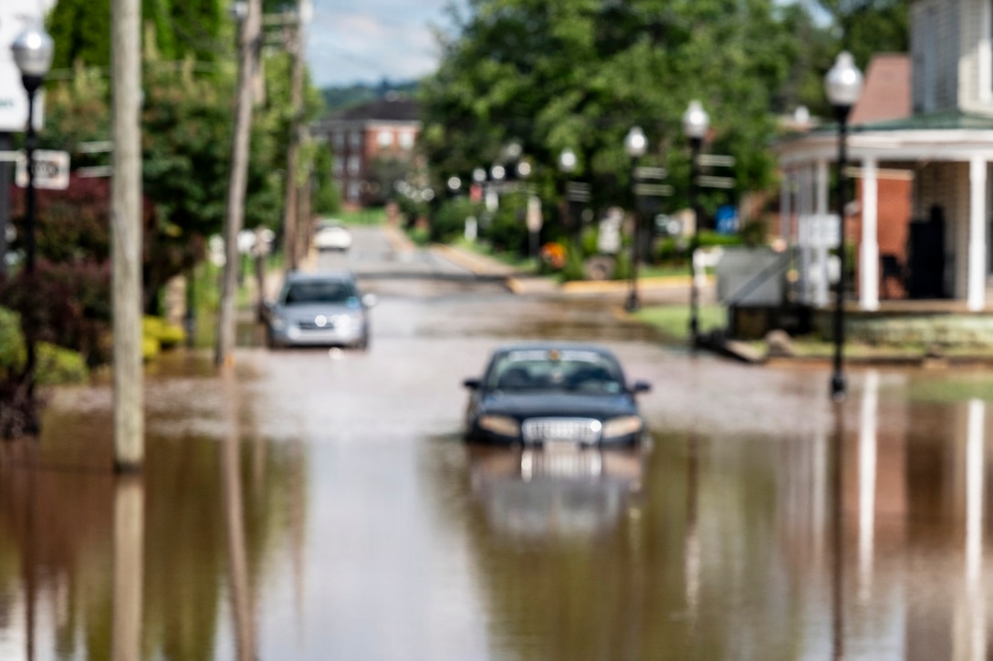A car is mostly submerged in deep water in the middle of a neighborhood street. A white barricade with a red sign that reads, "road closed," is in the forefront.